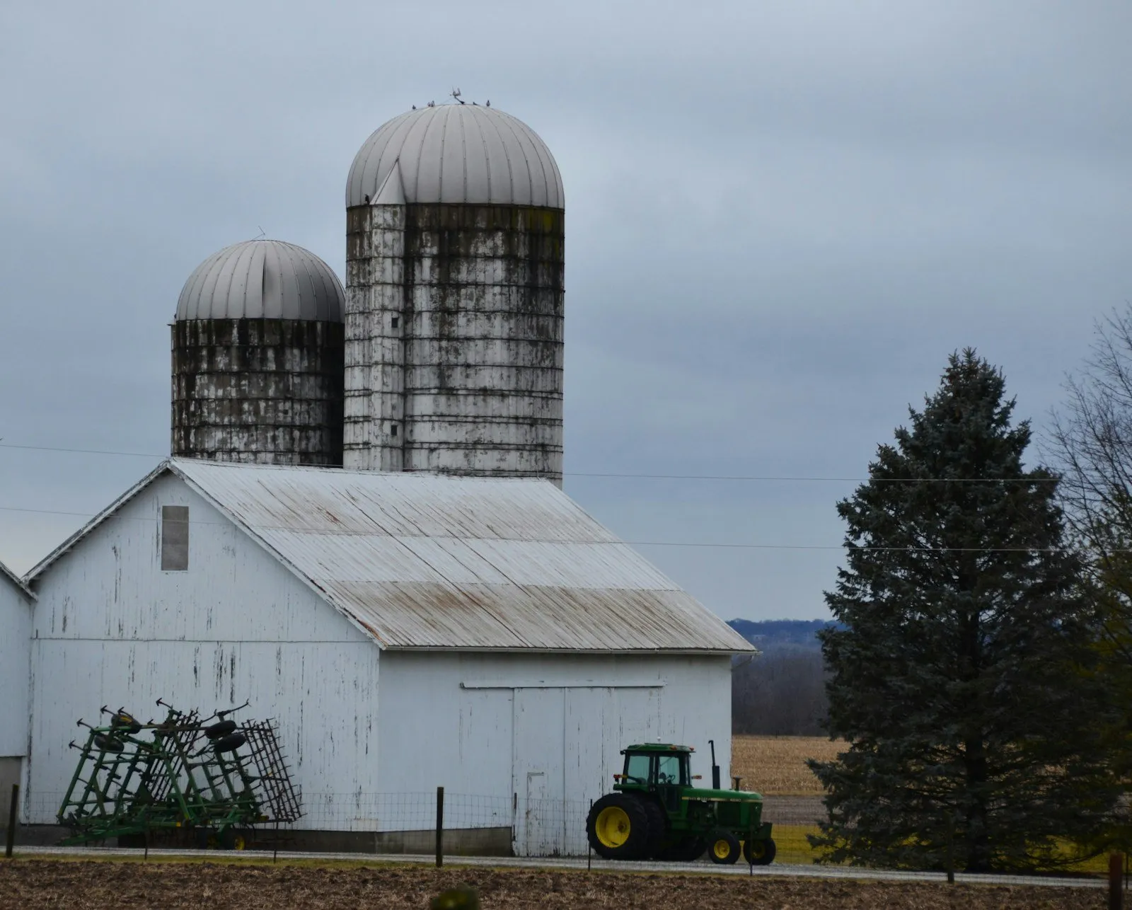 A white dairy barn with twin concrete silos, a green tractor parked beside it.